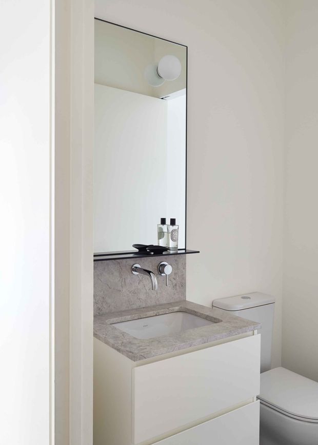 Compact contemporary bathroom viewed through a doorway, featuring a wall-mounted white vanity with integrated rectangular basin, pale stone countertop and matching short backsplash, wall-mounted chrome tapware, a large frameless mirror, and a white toilet. The room has a clean, restrained neutral palette and minimal detailing.