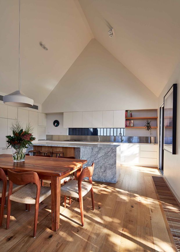 A bright, refined open-plan kitchen and dining area beneath a high pitched ceiling. The kitchen features white flat-panel cabinetry, open timber shelving, stainless-steel appliances and splashback details, and a prominent veined stone island. A solid timber dining table with woven-seat wooden chairs occupies the foreground. Warm sunlight enters through large glazing, highlighting the timber flooring and neutral interior.