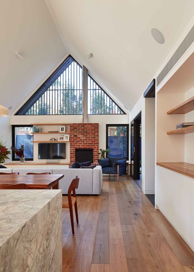 A bright open-plan interior combining a kitchen, dining and living area. The space features wide timber flooring, a large veined stone kitchen island, white built-in cabinetry, timber open shelving, a red brick fireplace wall, extensive black-framed glazing, and a dramatic triangular vaulted window beneath a high pitched ceiling. Mid-century-inspired timber dining furniture and upholstered seating add warmth.