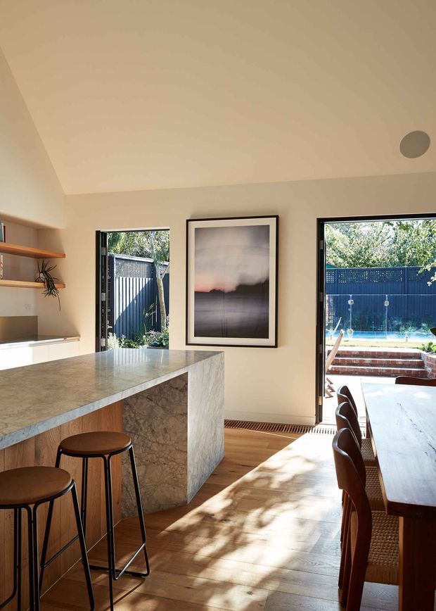 Bright contemporary open-plan kitchen and dining area with a large stone island, timber cabinetry and flooring, black-framed glazing, bar stools, and a dining table. The space opens directly to an outdoor pool courtyard through large doors, with warm natural light and neutral finishes.