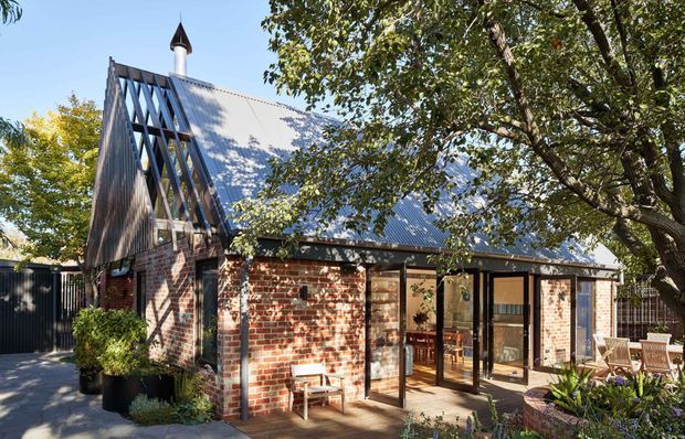 Exterior view of a distinctive brick building with a steep gabled corrugated metal roof, prominent exposed timber framing, and a small cupola. Large glazed doors open toward a paved outdoor terrace, with mature trees and planting surrounding the structure.