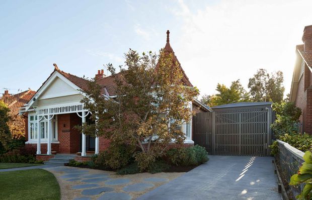Street-facing view of a character brick residence with ornate white timber verandah detailing, a steep gabled and turret-like roof form, landscaped front garden, stone stepping path, and a contemporary slatted garage door beside the home.