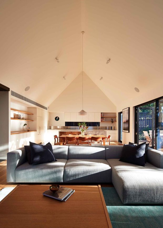A bright open-plan living and dining area with a kitchen at the rear. The room features a large grey sectional sofa, timber coffee table, built-in shelving and cabinetry, a timber dining table with upholstered chairs, and large glazed doors opening to an outdoor area. A dramatic pitched ceiling with a central pendant light creates a spacious, airy interior.