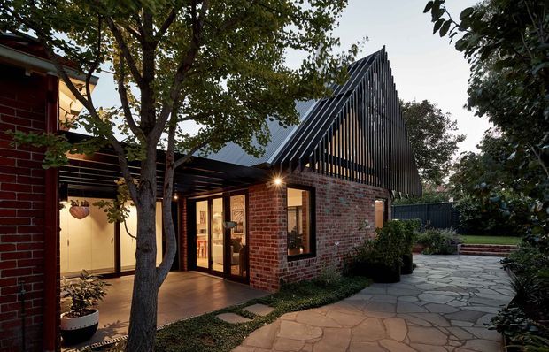 Exterior view of a contemporary brick house extension with a steep, sculptural gabled roof and prominent vertical dark metal fins. Warmly lit glazed doors open onto a paved garden courtyard, with established trees and planting surrounding the building.