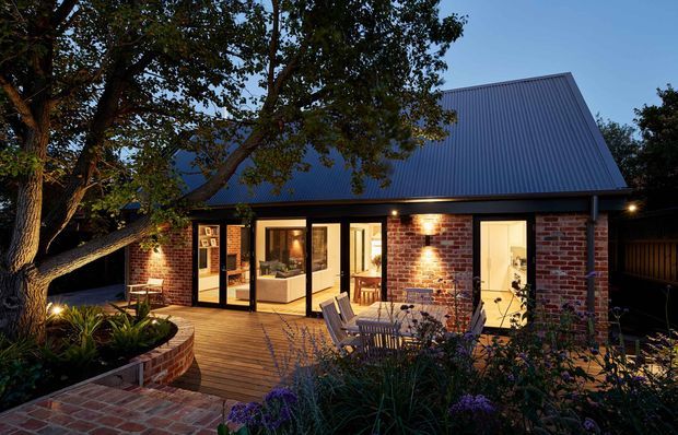 Twilight exterior view of a compact brick-clad home with a steeply pitched corrugated metal gable roof. The building opens onto a timber deck with outdoor dining furniture, while warm interior lighting glows through large glazed doors and windows. Mature trees, planted garden beds, brick paving, and curved brick retaining walls surround the dwelling.