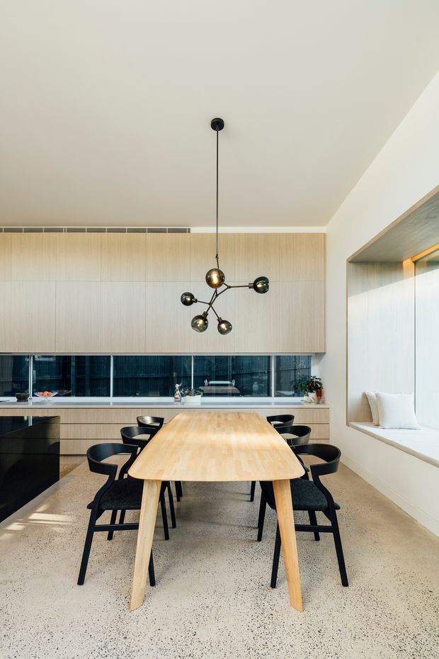 Contemporary dining area with a light timber dining table and black upholstered chairs, positioned beneath a sculptural multi-arm pendant light. The room features pale timber wall cabinetry, a long horizontal dark window or glazed opening, a light stone or terrazzo floor, and a built-in window seat with cushions.