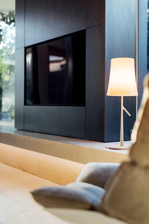 A contemporary living room interior featuring a dark wood built-in media wall with an integrated black television, a textured concrete ledge, and a slim floor lamp with a warm white shade. A neutral upholstered sofa and cushions occupy the foreground, with daylight visible through glazing at the side.