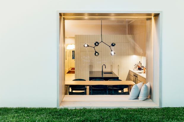 Exterior view looking through a large framed window into a contemporary dining and kitchen area. The interior features a timber dining table with dark chairs, a linear pendant light, pale vertical wall panelling, light cabinetry, and warm integrated lighting. A small glimpse of a living or hallway area is visible to the left, while cushions rest on the interior sill. The foreground includes grass outside.