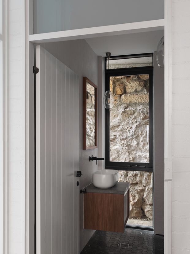 Compact contemporary bathroom with a wall-mounted timber vanity, white vessel basin, black tapware, and dark hexagonal floor tiles. White vertical wall panelling and painted brickwork frame a narrow black-framed window, which looks directly onto a textured natural stone wall. A narrow wall mirror and pendant light add functional detail.
