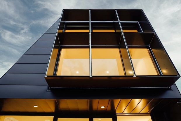 Low-angle exterior view of a modern multi-storey building facade with a projecting, box-like glazed volume. Dark metal cladding and framing contrast with warm amber-lit glazing and recessed downlights beneath the overhang; a cloudy sky is visible behind it.