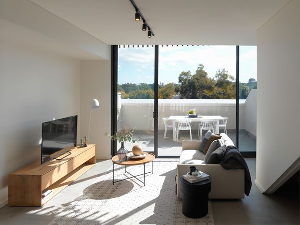 Bright contemporary living room with a low timber media console, television, neutral sofa, round coffee table, floor lamp, and patterned rug. Full-height sliding glass doors open onto a balcony terrace furnished with a white outdoor dining table and chairs. Track lighting, clean white walls, and strong afternoon sunlight create an airy, minimalist interior.