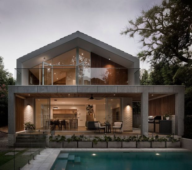 A contemporary two-storey residence viewed from the rear, with a strong pitched gable roof, concrete-framed upper terrace, extensive glazing, and warm vertical timber cladding. The ground floor opens to an outdoor entertaining terrace beside a rectangular swimming pool, with an illuminated kitchen, dining, and living area visible through the glass. Built-in planters and mature trees frame the landscaped setting.