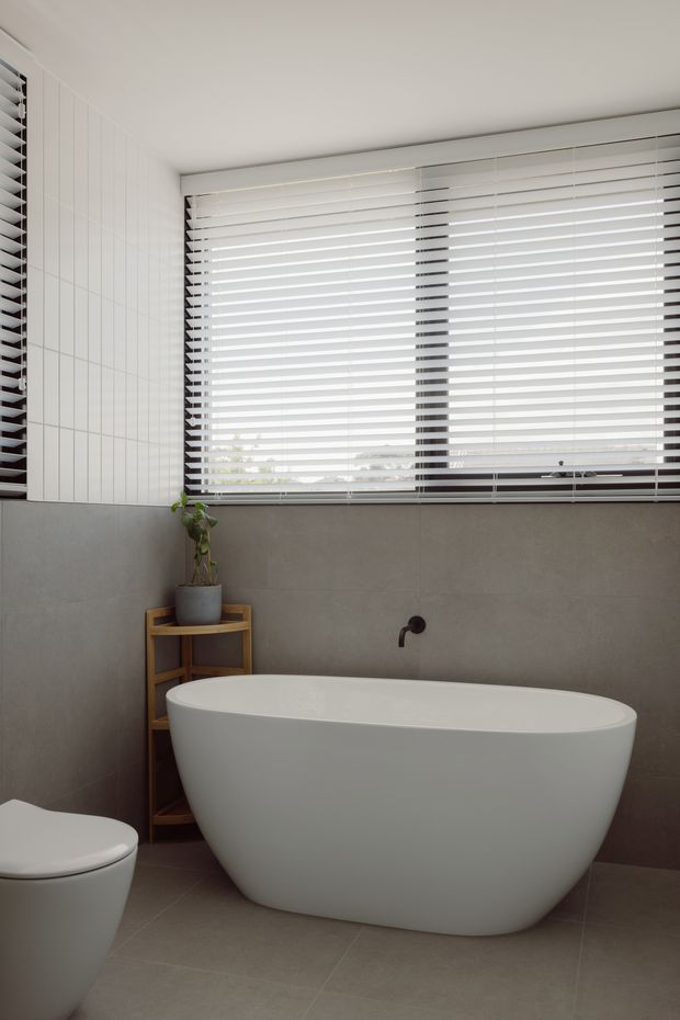 Bright contemporary bathroom featuring a freestanding white bathtub, partially visible toilet, large horizontal window with venetian blinds, grey tiled walls and floor, white vertical wall tiles, and dark wall-mounted tapware. A small timber plant stand adds warmth.