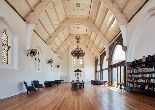 A grand, elongated hall with a steeply vaulted timber-lined ceiling, white walls, ornate chandeliers, tall arched windows, and polished timber flooring. Dark built-in bookshelves line one side, while scattered lounge and dining furniture occupy the spacious interior. Gothic-inspired arched detailing, stained-glass windows, exposed decorative structure, and abundant natural light create a formal yet inviting atmosphere.