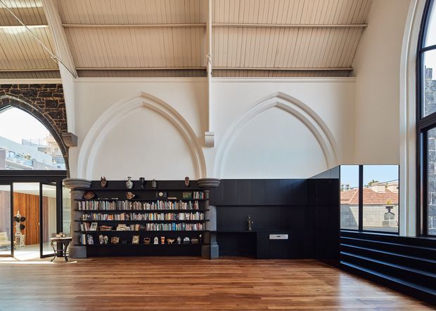 A dramatic adaptive interior with a tall, white vaulted ceiling, prominent pointed-arch wall recesses, exposed dark stone masonry, and large arched windows. Warm timber flooring contrasts with a substantial black built-in library and media/storage joinery, creating a refined blend of historic architectural detailing and contemporary fit-out.