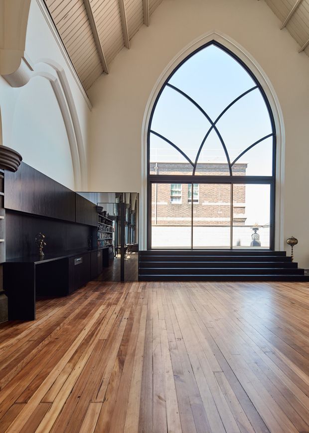 A tall, light-filled interior with a dramatic arched steel-framed window, exposed pitched timber ceiling, warm timber plank flooring, and dark built-in cabinetry along one wall. A stepped platform sits beneath the window, while the reflected cabinetry and shelving extend the sense of depth. The space has a refined, gallery-like character blending historic architectural detailing with contemporary joinery.