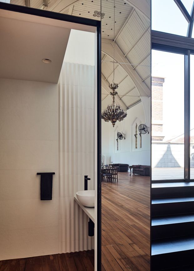 A contemporary bathroom is shown in the foreground, with pale textured wall surfaces, a white vessel basin on a slim vanity, matte black tapware and towel rail, and dark timber flooring. Full-height black-framed glazing and a mirrored/reflected view open onto an adjoining grand high-ceilinged hall or living space with exposed white-painted rafters, timber flooring, ornate chandeliers, arched wall detailing and dark-framed stair elements.