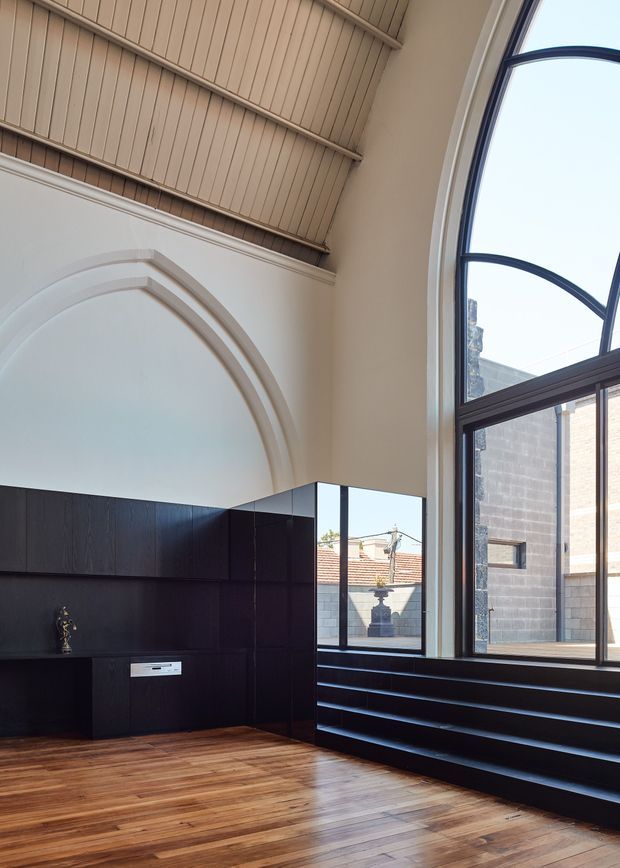 A tall, refined interior with a dramatic arched window and large arched wall recess, pale plaster walls, a timber-lined vaulted ceiling, dark built-in cabinetry, stepped window seating or display shelving, and warm timber flooring. The space appears to be a living or gallery-like room, with strong contrast between the dark joinery and light architectural envelope.