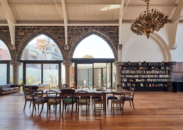 A spacious, high-ceilinged interior combining historic Gothic-inspired masonry with a contemporary library and dining area. Large pointed-arch openings are framed by dark textured stone and slender columns, while an ornate chandelier hangs above timber dining tables and mixed vintage-style chairs. A full-height black bookshelf occupies the right wall, and tall glazed doors and windows bring in abundant daylight. The room has polished warm timber flooring and a refined heritage-meets-contemporary atmosphere.