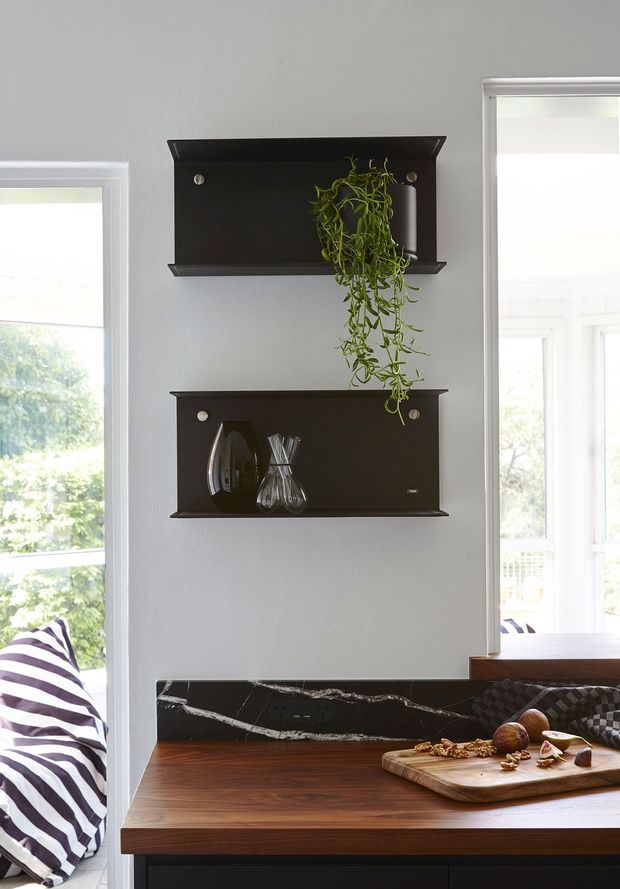 Bright interior vignette showing a compact kitchen or dining nook with dark floating display shelves, a trailing plant, decorative glassware, a dark marble-look splashback, and a warm timber countertop. Large windows provide abundant natural light, with a striped upholstered bench visible at left.