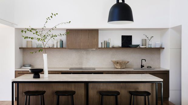 A refined contemporary kitchen viewed straight on, featuring a large island with dark timber cabinetry, a pale stone benchtop, four black stools, and a minimalist black pendant light. The rear wall has matching timber cabinetry, open timber shelving with ceramics and greenery, a pale textured splashback, and integrated cooking and sink areas. The palette is warm and neutral with black accents.