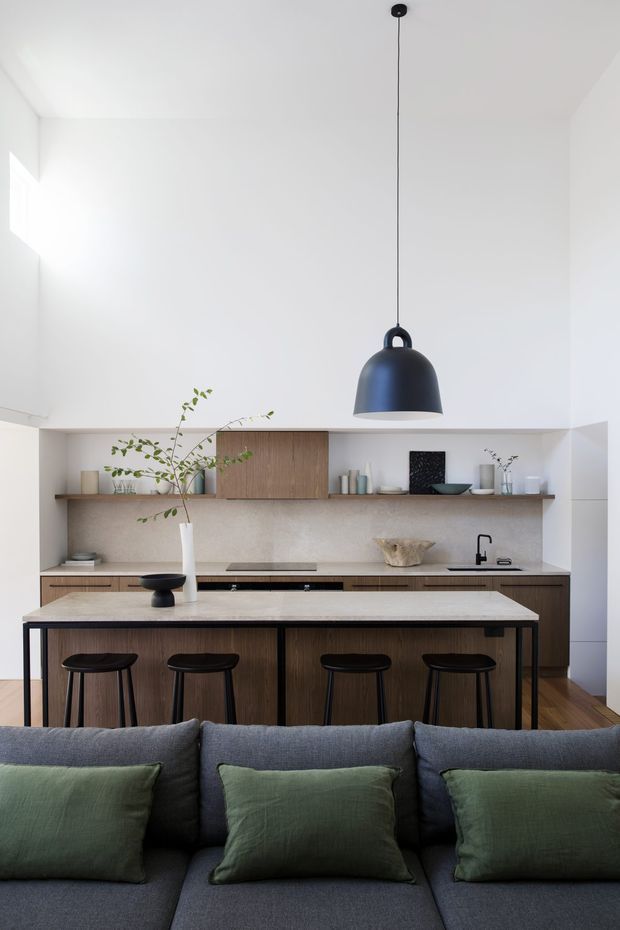 A bright, minimalist open-plan kitchen viewed from behind a dark grey sectional sofa. The kitchen features warm timber cabinetry, a pale stone island benchtop with four black stools, open shelving, a light neutral splashback, integrated appliances, and a large matte black pendant light suspended from a high white ceiling. Green cushions and a leafy branch add muted natural accents.