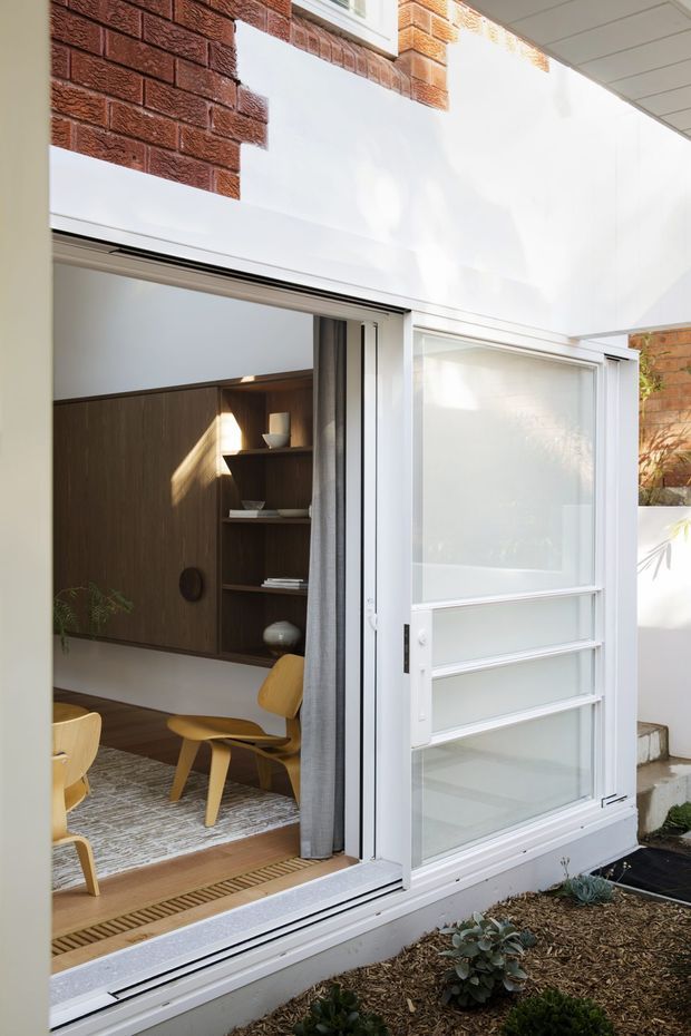 A bright architectural view from a small planted courtyard into a contemporary dining and living area through large white-framed glazed sliding doors. The interior features a warm timber built-in wall with open shelving, light timber dining chairs and a low table, while exposed red brick walls, white plaster surfaces and a translucent glazed door define the courtyard edge.
