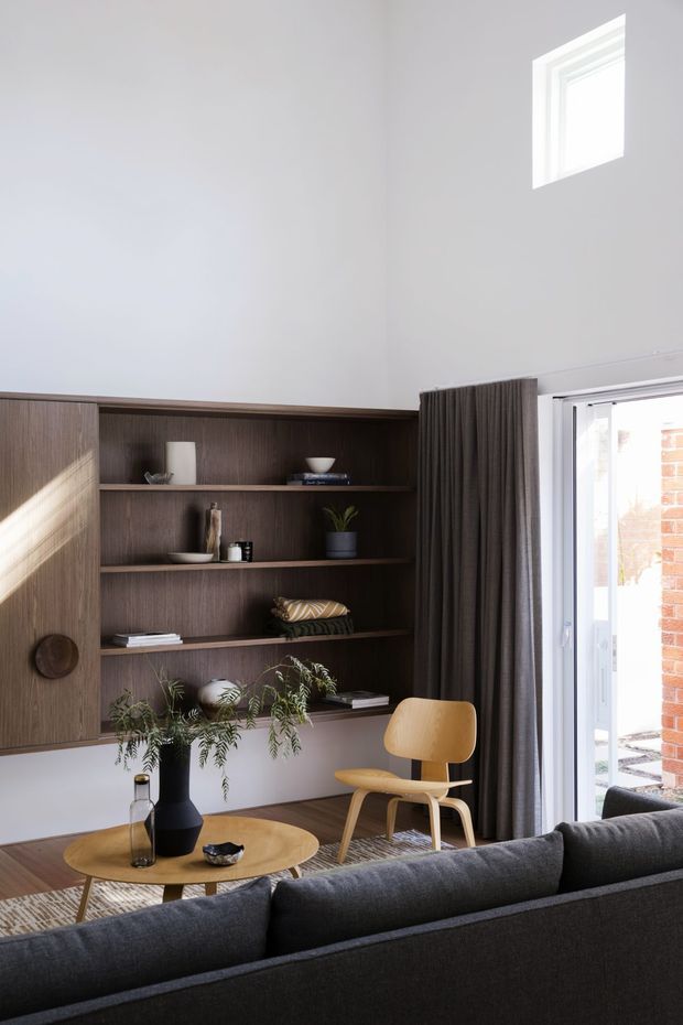 A bright, compact living room with a tall white ceiling and white walls. A dark walnut-toned built-in shelving and storage unit spans the left wall, displaying books and decorative objects. A grey upholstered sofa faces a low round timber coffee table, with a sculptural light-wood armchair beside it. Full-height dark curtains frame glazed exterior doors, and a small high-set window brings in additional daylight. Indoor plants and a neutral textured rug add warmth and softness.