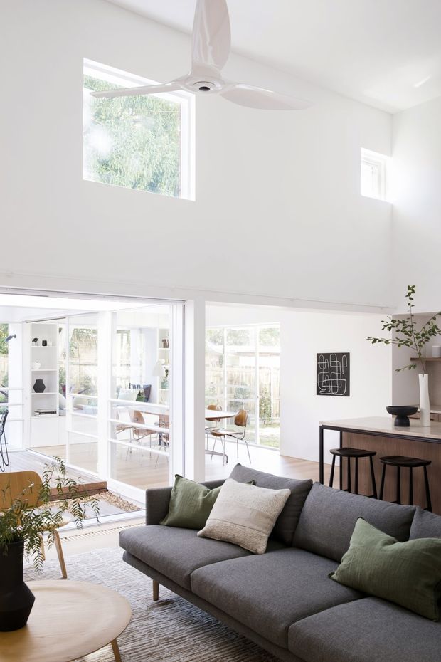Bright open-plan living area with a large grey sectional sofa in the foreground, light wood coffee table, pale textured rug, and soft green and cream cushions. A compact kitchen with dark lower cabinetry, a pale benchtop and bar stools sits to the right, while extensive glazing connects to a light-filled dining or sunroom area. The room has white walls, a high ceiling with a white ceiling fan, and abundant natural light.