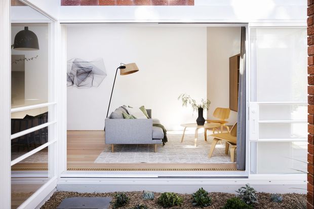 A bright, minimalist living room viewed from a small landscaped courtyard through wide-open glass doors. The room features pale timber flooring, a light grey sofa with cushions, a neutral patterned rug, a black floor lamp with a timber shade, sculptural wall art, a small timber coffee table, and two mustard-yellow lounge chairs. White built-in shelving and door frames surround the opening, with exposed red brick visible at the exterior edge.