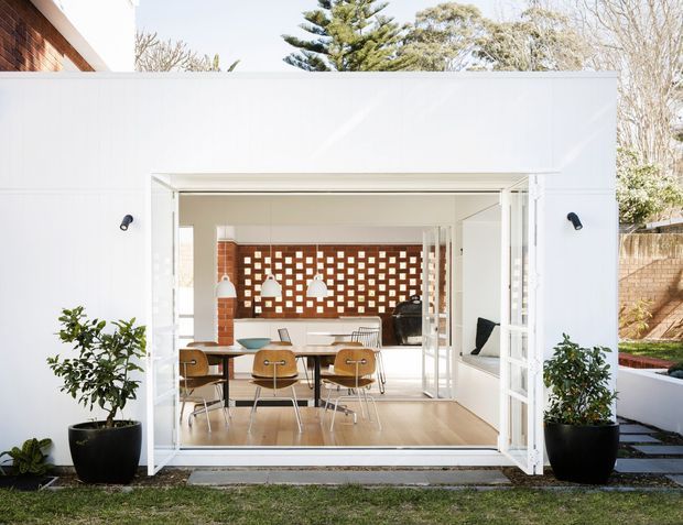 Bright exterior view into a contemporary indoor-outdoor living and dining space through fully opened white bi-fold doors. The room features a light timber floor, dining table with chairs, a white kitchen or serving area, and a distinctive terracotta brick breeze-block screen at the rear. White rendered walls, black wall lights, potted plants, and a small garden terrace frame the opening.