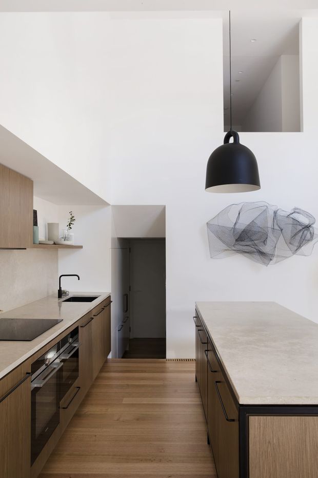 A refined contemporary kitchen with a compact galley layout and large central island. The space features warm timber cabinetry, pale stone benchtops, integrated appliances, a black undermount sink tap, and a matte black pendant light. White walls and a high open ceiling create a bright, minimal interior, with a sculptural wire artwork mounted on the wall.