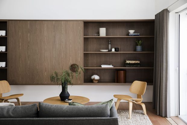 A refined contemporary living room viewed from behind a dark grey sofa, featuring a full-width dark timber built-in wall unit with concealed cabinetry and open display shelving. The room includes a low round timber coffee table, sculptural timber lounge chairs, a neutral textured rug, indoor plants, and full-height glazing dressed with dark curtains. The palette is warm and earthy with clean-lined, understated styling.
