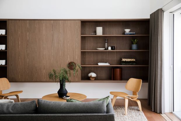 A refined living room with a built-in dark timber wall unit combining concealed cabinetry and open display shelving. A grey sofa faces a round timber coffee table, while two light timber lounge chairs sit near large glazed doors or windows with full-height grey curtains. The room has white walls, warm timber flooring, restrained decorative objects, and abundant natural light.