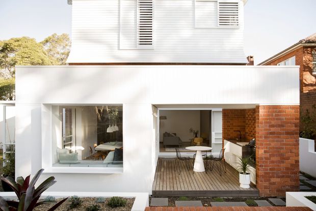 Front exterior view of a contemporary two-storey residence with a crisp white rendered lower volume, white horizontal cladding and louvred window shutters above, plus a contrasting red brick wall. Large glazed openings reveal a living area and dining space, while a timber-decked recessed outdoor area and landscaped front garden sit in the foreground.