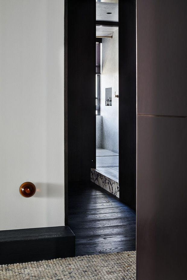 A narrow view through a partially open dark timber doorway into a refined bathroom. The bathroom features pale small-format tiled walls, a tiled shower area with dark fixtures, and a marble-clad bath or ledge with strong veining. The foreground has dark timber flooring, dark built-in joinery or door panels, a white wall with a small rounded timber handle, and a textured neutral wool rug. The composition is intimate and architectural, emphasizing layered thresholds, contrasting dark wood, pale tile, and natural stone.