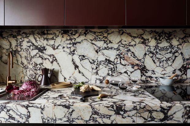 A close-up view of a richly detailed kitchen featuring dramatic veined marble across the splashback, benchtop, and island cabinetry fronts. Dark burgundy cabinetry runs along the upper wall, while a brass-toned tap, dark sink, kettle, crockery, wine glasses, and food styling add warmth and a sophisticated, luxurious character.