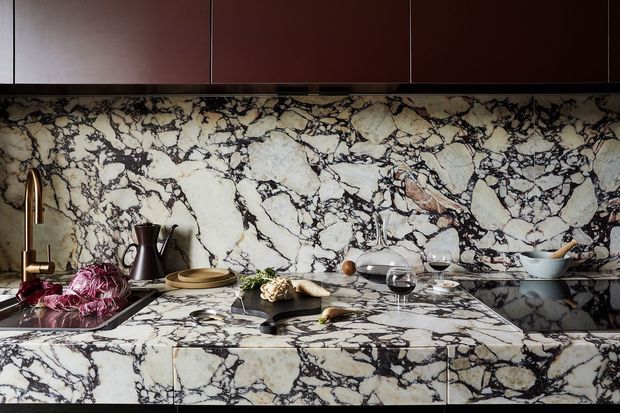 Close-up view of a dramatic kitchen with extensive veined stone surfaces. A dark timber-toned wall of upper cabinetry sits above a matching stone splashback, benchtop, and waterfall-style island or peninsula. The richly patterned pale stone has bold black and warm rust veining. A sink, brass-toned tapware, dark kettle, cookware, dishes, and wine glasses are visible, creating a high-end, richly layered kitchen composition.