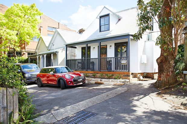 Bright street-level exterior view of a compact white weatherboard dwelling with a steep gabled corrugated roof, dormer window, covered front verandah and dark timber railing. The house sits on a narrow urban site with a concrete driveway, mature trees, and neighbouring buildings visible around it.