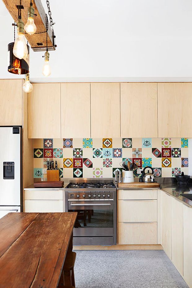 Bright, compact kitchen with pale timber flat-panel cabinetry, a stainless-steel range cooker and refrigerator, dark benchtops, and a colourful patchwork patterned tile splashback. A rustic timber dining table and hanging exposed-bulb pendant lights add warmth, while the speckled grey floor and white walls keep the space light.