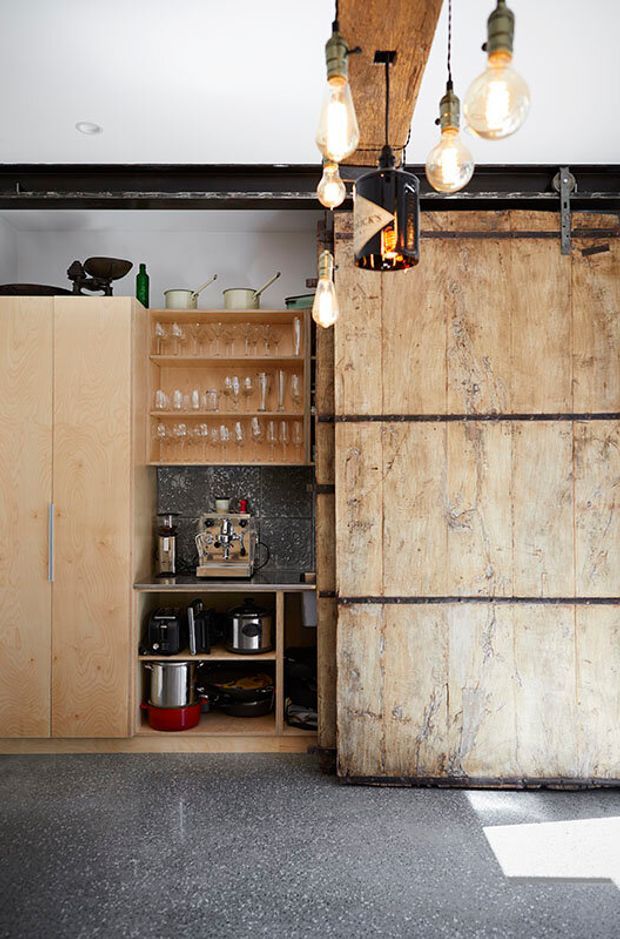 A compact industrial-style kitchen or bar area with pale timber cabinetry, open shelving displaying glassware, a coffee machine and small appliances, and a large weathered timber sliding barn door. Exposed dark metal trackwork and hanging vintage-style filament lights add an industrial character. The floor is a dark speckled polished surface.
