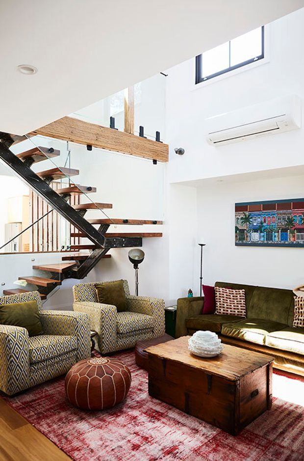 A bright, double-height living room with a dramatic open-riser timber and black steel staircase. White walls, clerestory glazing, and an exposed timber landing create an airy contemporary shell. The room is furnished with patterned armchairs, an olive-green sofa, a rustic timber coffee table, a red patterned rug, and a round leather ottoman.