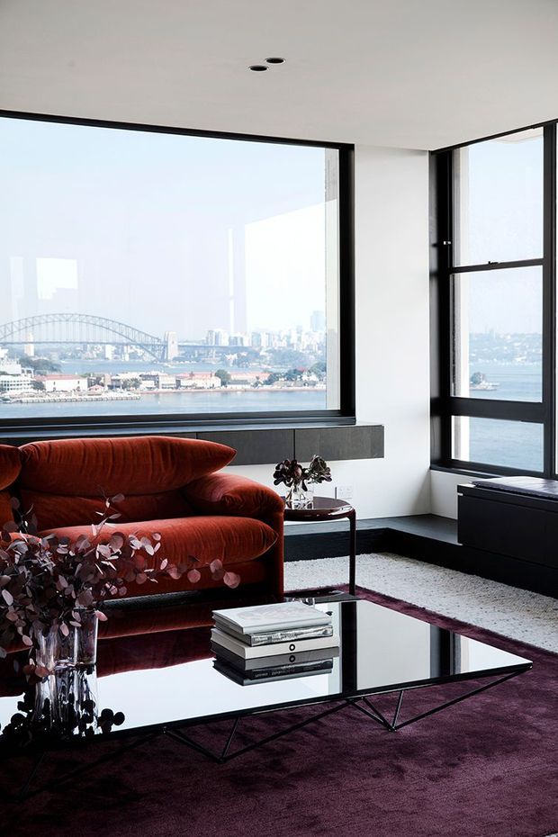 A sophisticated living room with expansive black-framed corner glazing overlooking a harbour and city skyline. The room features a sculptural rust-red sofa, black reflective geometric coffee table, dark purple carpet, white walls and ceiling, and a low dark built-in window seat or cabinet. The composition is contemporary and highly polished, with strong indoor-outdoor visual connection and dramatic contrast.