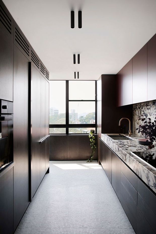 A narrow, contemporary galley kitchen with full-height dark timber or stained cabinetry on both sides, integrated appliances, black-framed floor-to-ceiling windows at the end, and a dramatic veined marble benchtop and splashback. Minimal black cylindrical ceiling lights, a dark gooseneck tap, and a small indoor plant complete the refined, high-contrast interior. The space is brightened by daylight and offers an elevated urban outlook.