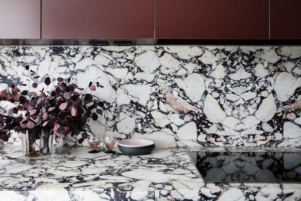 Close-up view of a sophisticated kitchen or bar area featuring dramatic white marble with dense dark veining across the splashback and benchtop. Dark burgundy-brown flat-panel cabinetry runs above the stone, while a vase of deep purple foliage and small decorative vessels sit on the counter. A dark inset sink or cooktop is visible at right.
