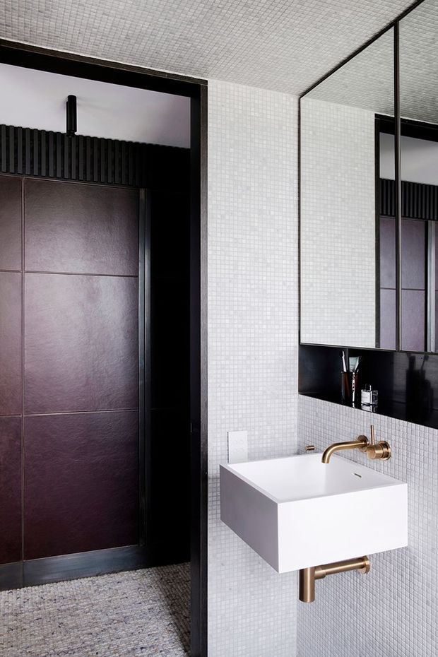 A compact, highly detailed contemporary bathroom with white small-format mosaic tiles covering the walls and ceiling. A wall-mounted rectangular white basin sits beneath a large frameless mirror, paired with exposed brushed brass plumbing and tapware. The adjacent dark doorway and wall panels feature black ribbed detailing and deep mauve-brown slab panels, creating a strong monochromatic contrast. The floor appears to be a textured grey stone or tile finish.