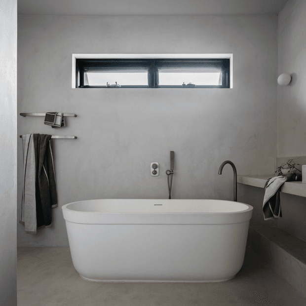 A minimalist bathroom with a freestanding white bathtub centered beneath a narrow horizontal window. The room has light grey plastered walls, a grey concrete-look floor, dark metal tapware, simple towel rails with neutral towels, and a raised ledge with a basin tap at right.