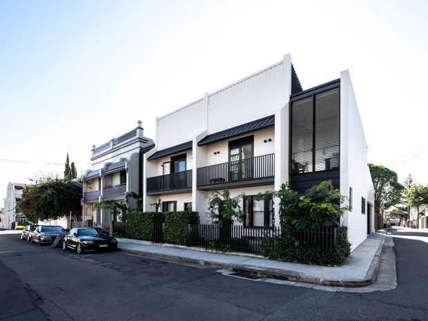 Street-level exterior view of a contemporary multi-storey residential building in an established urban neighbourhood. The facade combines white rendered walls, dark metal balcony railings, vertical screening and a pitched roof section, with landscaped planting along the frontage. An older ornate neighbouring terrace-style dwelling is visible beside it.