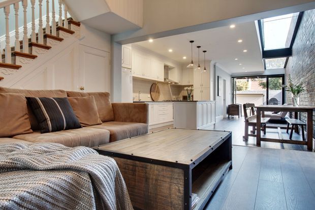 Bright open-plan living and dining space with a visible kitchen, featuring a large tan sectional sofa, industrial-style timber and metal coffee table, white shaker-style cabinetry, pendant lights, and a dining area extending toward full-height glazing. A staircase with turned balusters and timber handrail adds a traditional architectural detail, while a roof window brings in additional natural light.