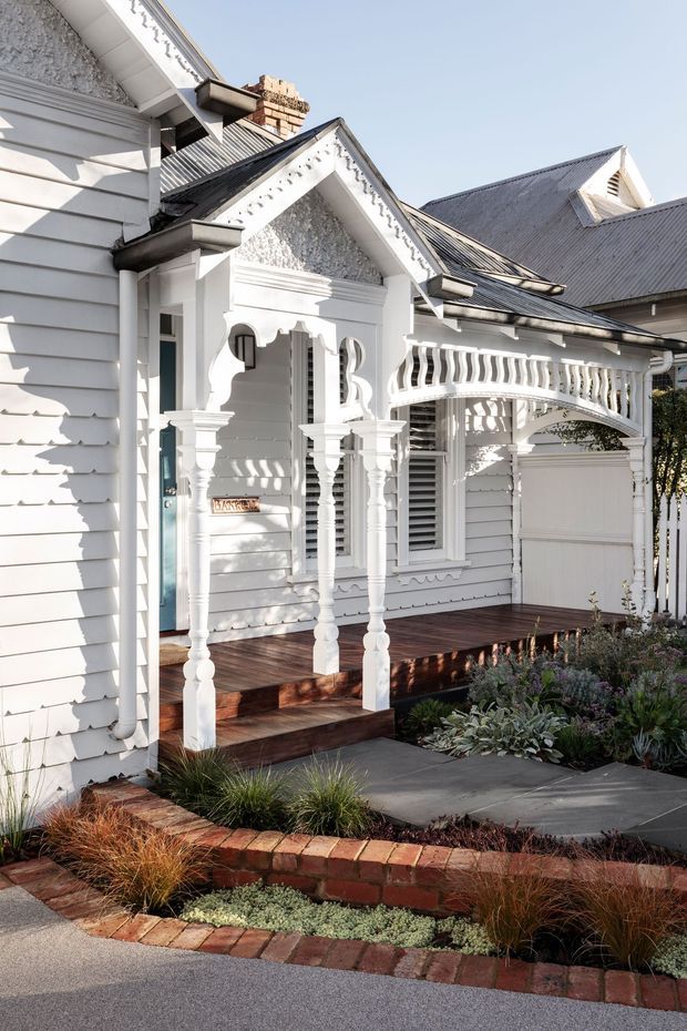 A bright front exterior view of a traditional white weatherboard house with ornate decorative timber verandah posts, scalloped trim, gabled roof sections, corrugated metal roofing, and a curved decorative carport or garage entrance. The home has a dark timber porch deck, white louvred windows, a turquoise front door, and a landscaped garden with brick edging and drought-tolerant planting.